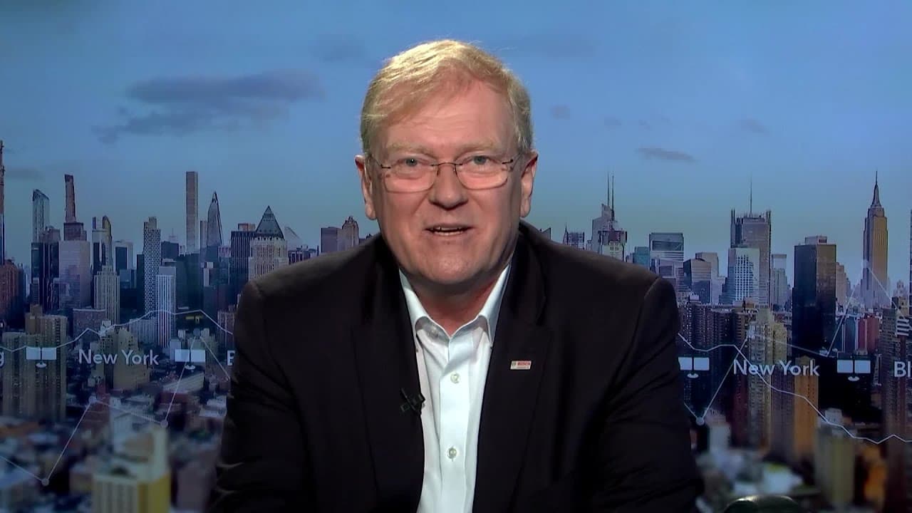 Two men in discussion, one on the left in a suit and glasses, the other on the right in a suit, with a cityscape background.