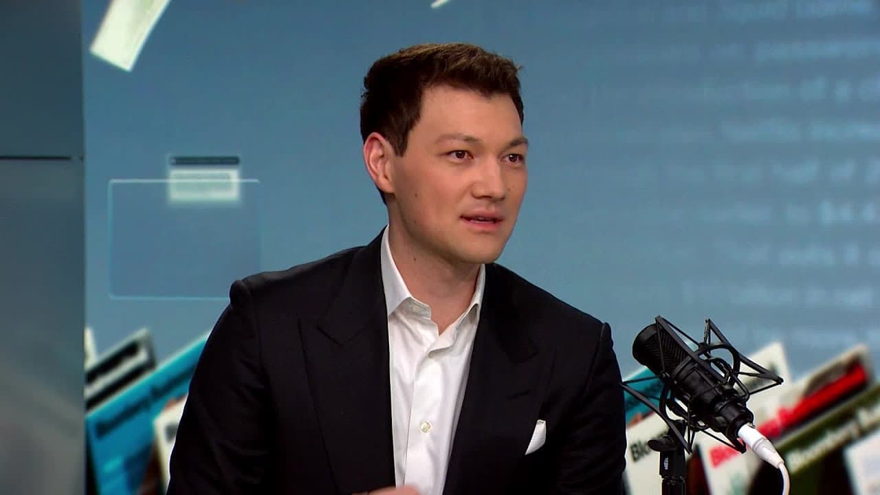 A man in a suit speaking on a broadcast, with a microphone in front of him.